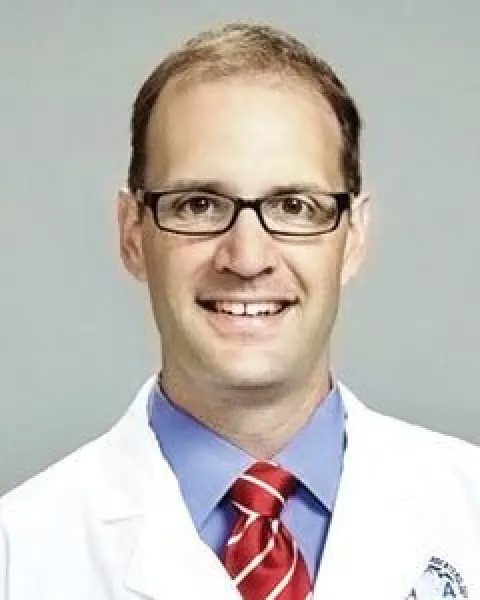 Charles W. Shrode, MD, smiling, wearing glasses, a blue shirt, red striped tie, and white lab coat posed against a neutral background.