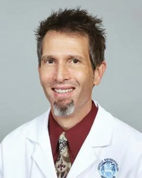 Craig J. Cender, MD, smiling in a white lab coat with a maroon shirt and patterned tie, against a gray background.